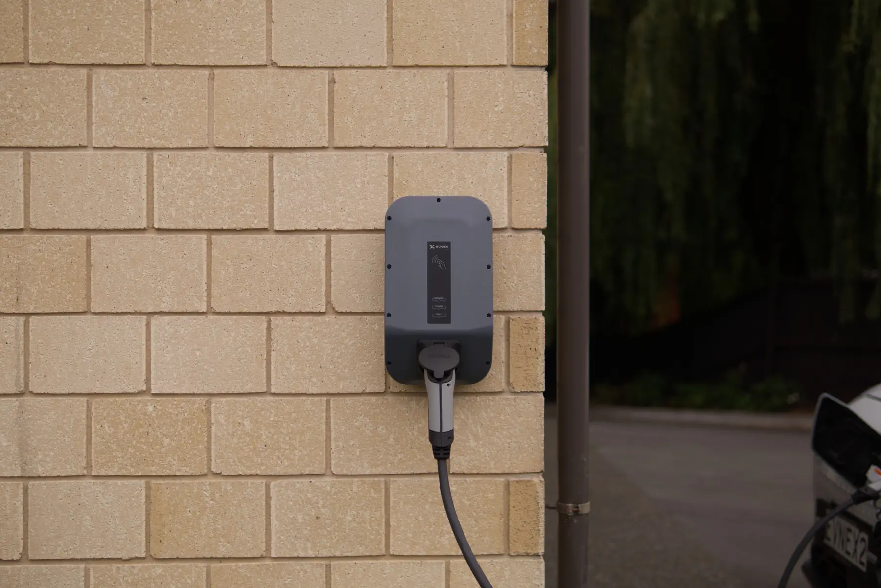 Een grijze thuislader voor elektrische auto's op een lichte bakstenen muur, met de kabel naar beneden