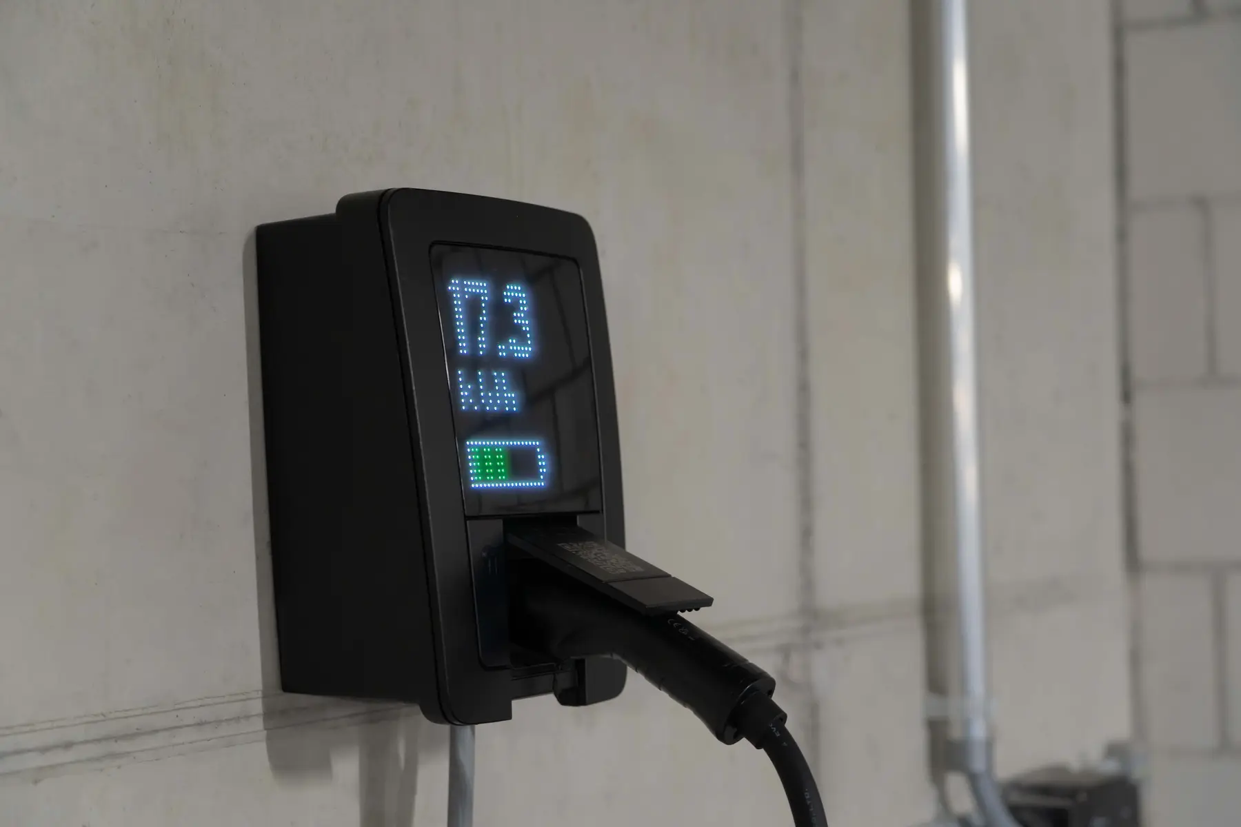 Een zwarte laadpaal aan de muur met verlicht display en een aangesloten laadkabel